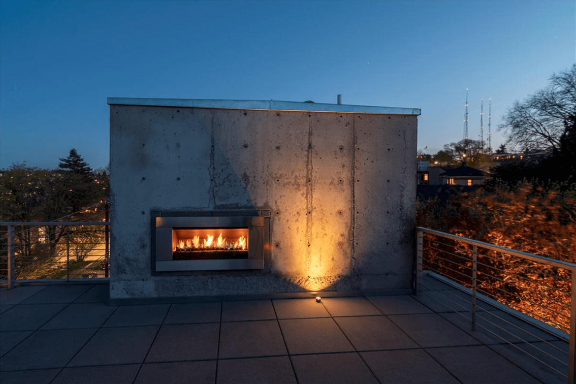 Architectural uplighting accentuates the concrete fireplace wall on a rooftop terrace overlooking Seattle at dusk. Northwest Outdoor Lighting
