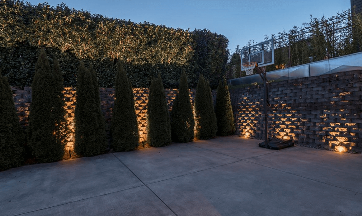 Warm landscape uplighting highlights evergreen hedges and a textured masonry privacy wall in the outdoor sport court of a Capitol Hill home in Seattle. Outdoor lighting designed and installed by Northwest Outdoor Lighting. Photography copyright © Northwest Outdoor Lighting. All rights reserved.