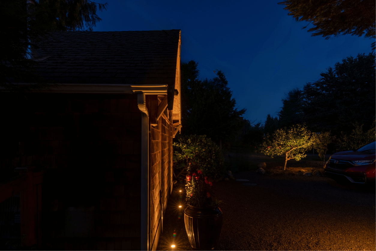 Side view of a Woodinville cottage garage illuminated with 18 Sunset Tone landscape lights, softly lighting the entry path, siding, and surrounding trees.