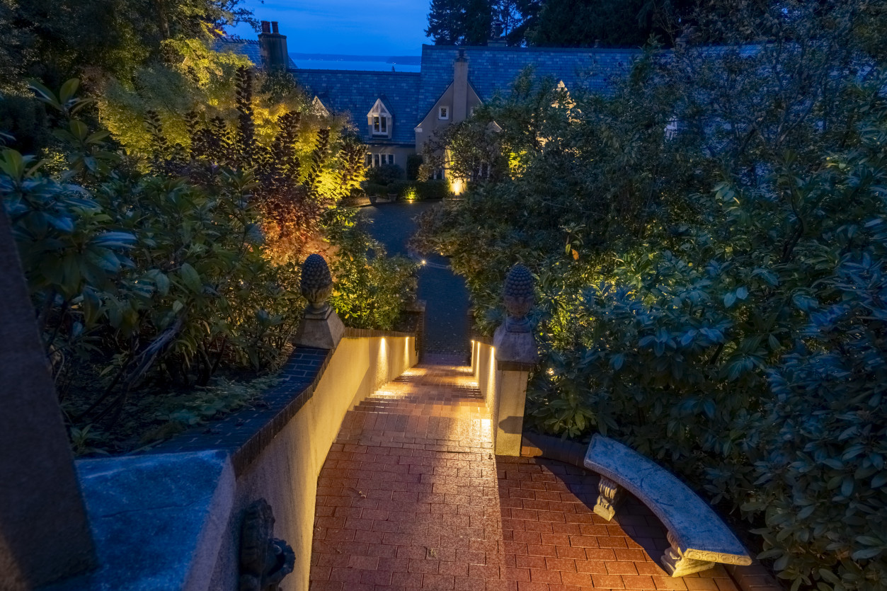 Brick pathway and stairway lighting leading to the driveway with warm LED fixtures highlighting greenery and retaining walls.