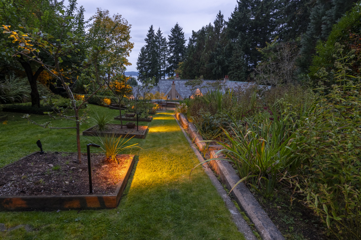 Garden walkway featuring low-voltage path lights leading across lawn and raised beds toward surrounding evergreen landscape.