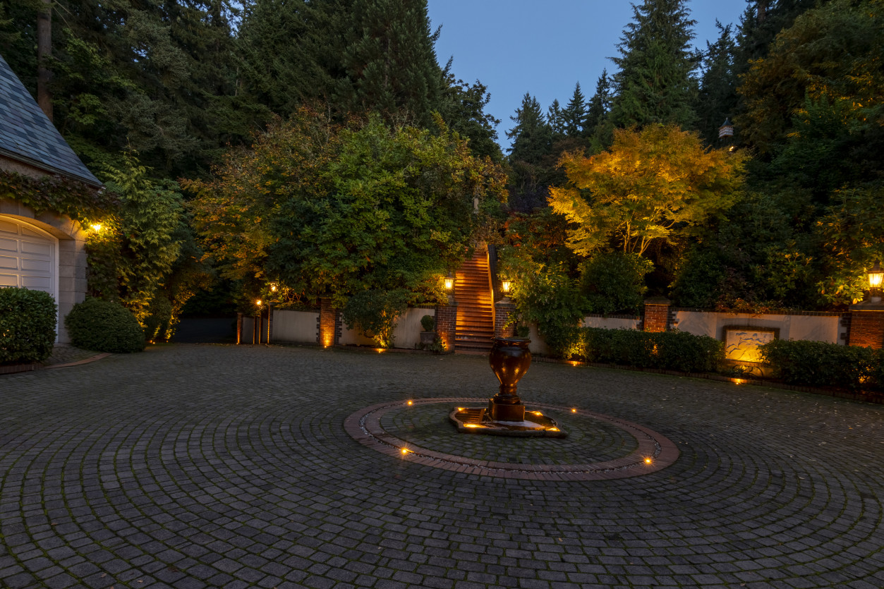 Illuminated cobblestone circular driveway with center fountain and surrounding landscape lighting at The Highlands Residence.