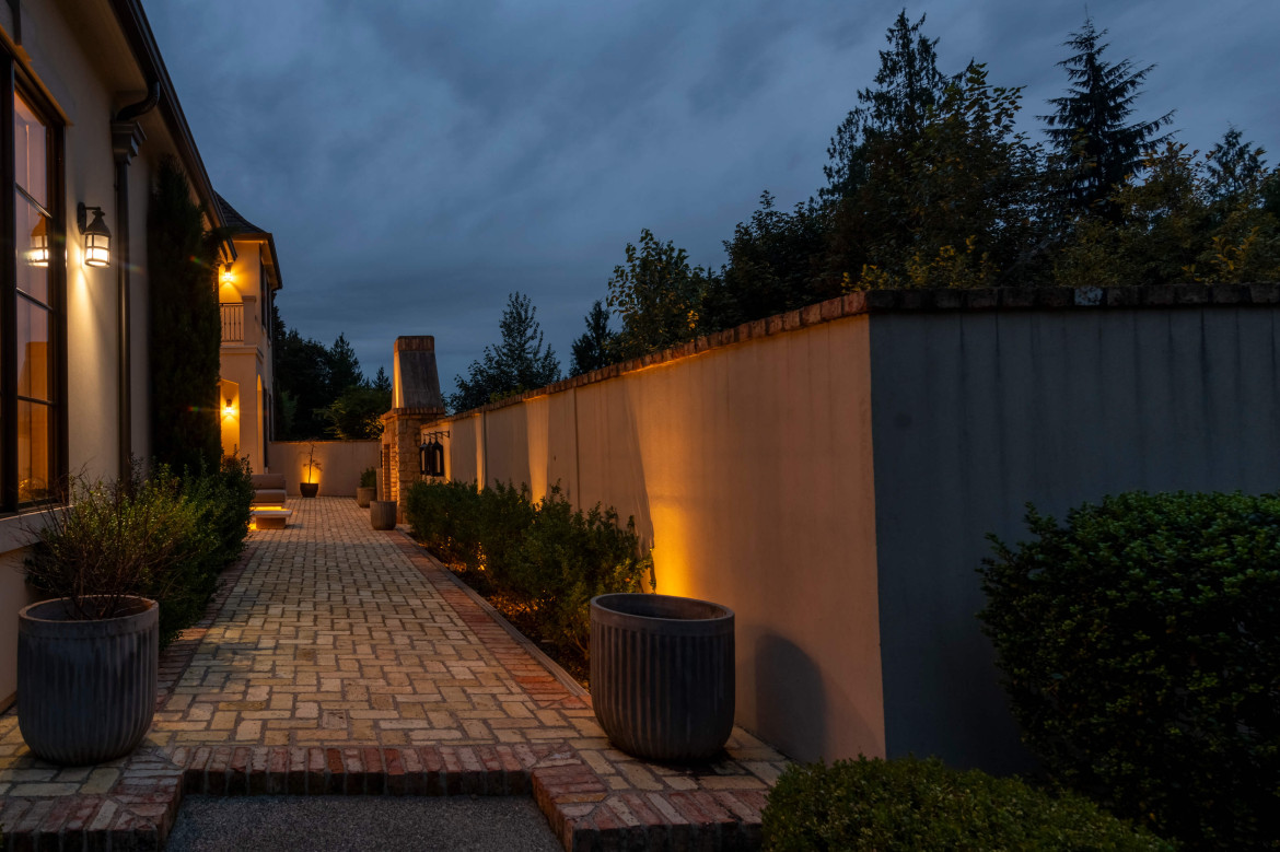 Warm wall-washing lights illuminate a long stucco privacy wall while landscape uplighting highlights tall evergreen trees along the driveway at Issaquah Manor in Issaquah, Washington. Outdoor lighting designed and installed by Northwest Outdoor Lighting. Photography copyright © Northwest Outdoor Lighting. All rights reserved.