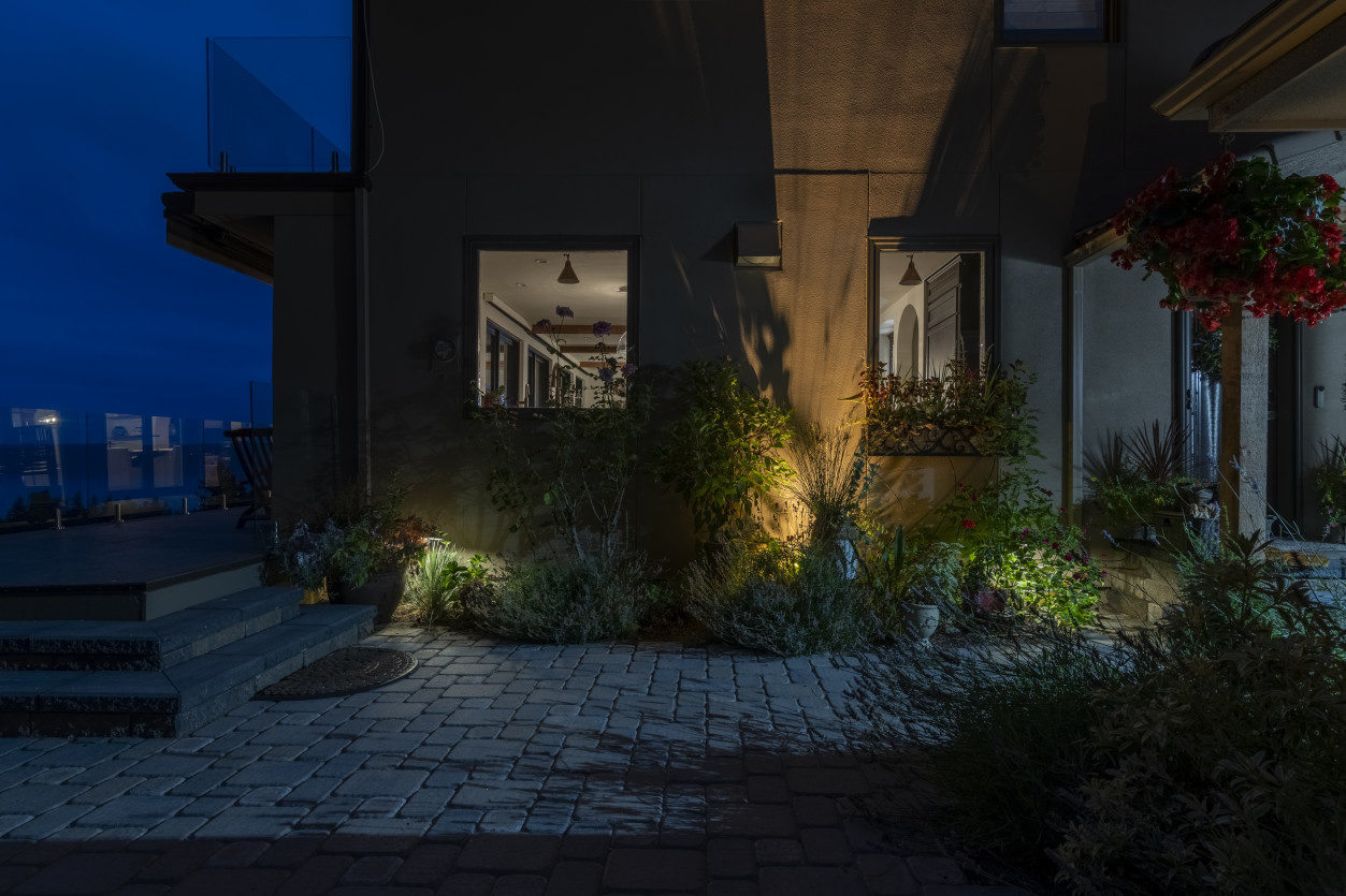 Architectural lighting highlighting stone façade and planting beds along the patio of the main residence at night.