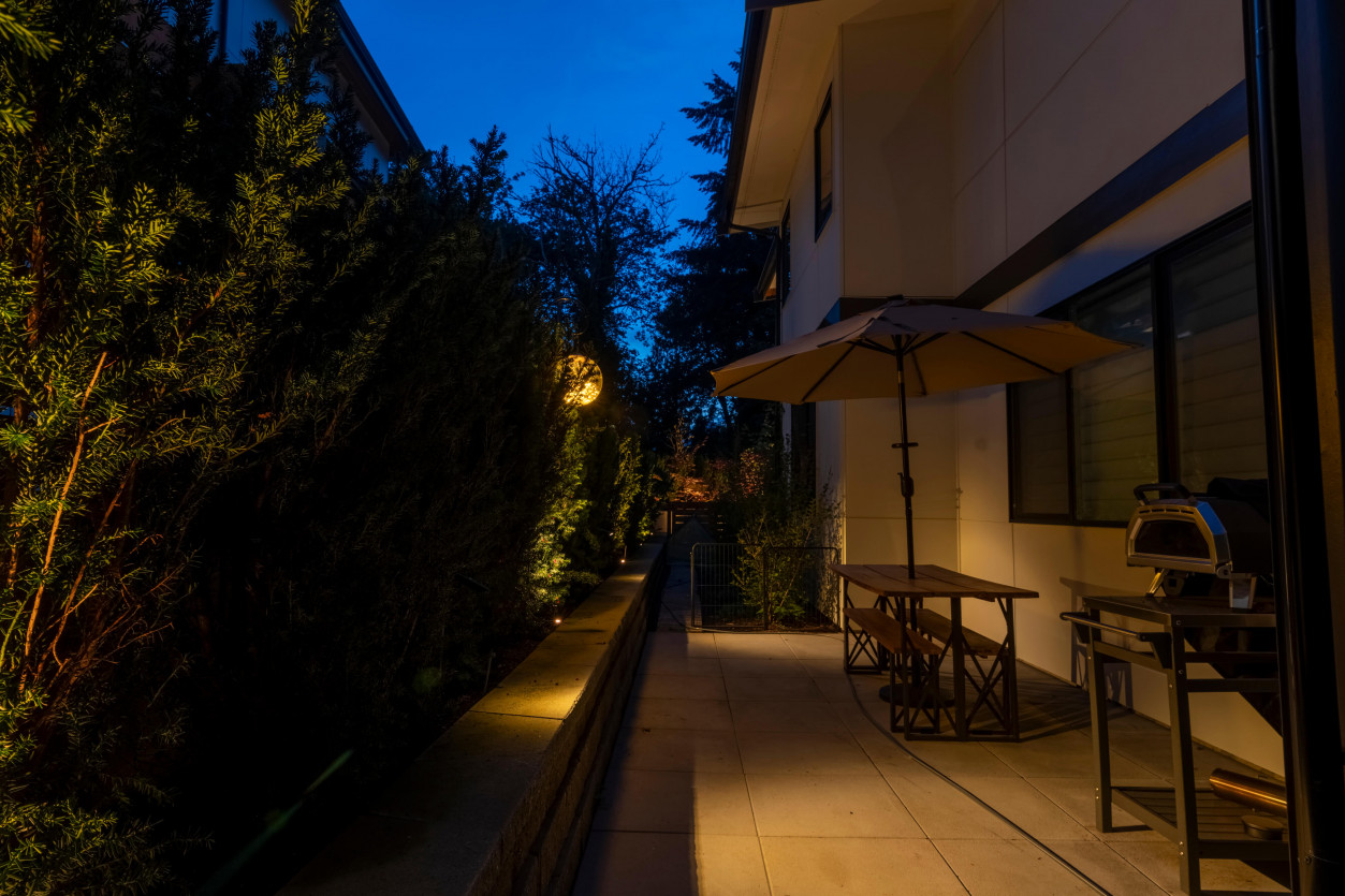 Side walkway and patio lighting softly illuminating pathway alongside the home with Sunset Tone fixtures and surrounding plantings.