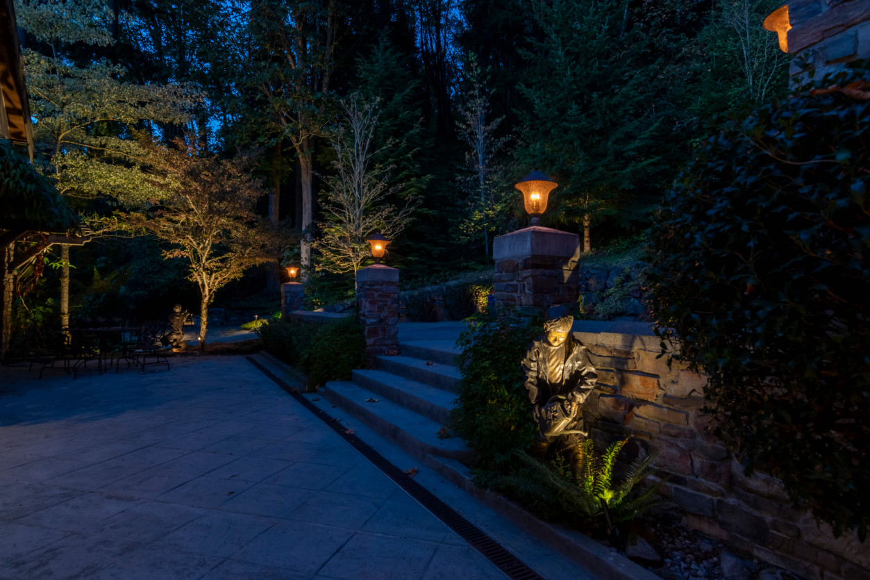 Nighttime driveway view with layered uplighting on tall evergreens and pathway markers at luxury Sammamish property.