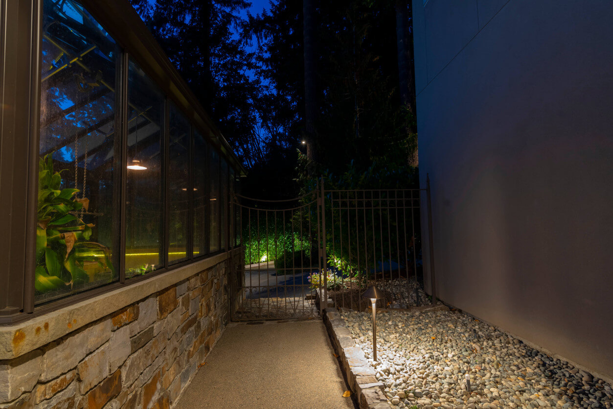 Soft pathway lighting leading to a glass conservatory greenhouse, illuminating stone foundation and modern exterior at a Sammamish estate designed by Northwest Outdoor Lighting.