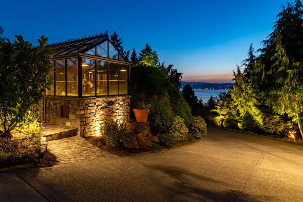 Glass garden conservatory glowing with warm lighting beside illuminated potted plants and stone terrace at a Sammamish estate.