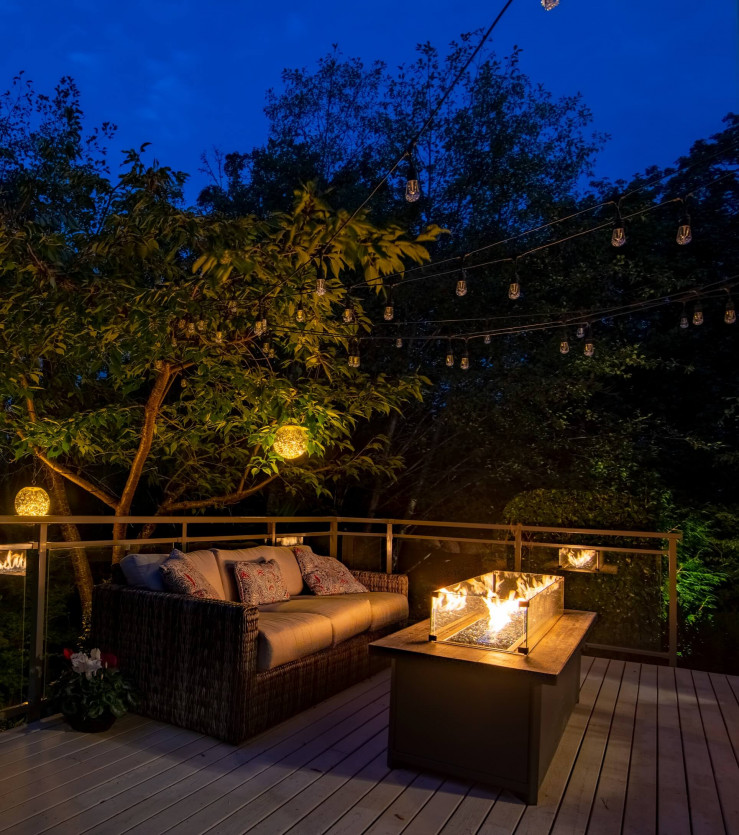 Firepit area glowing with 2700K lighting on upper deck lounge space with surrounding forest.