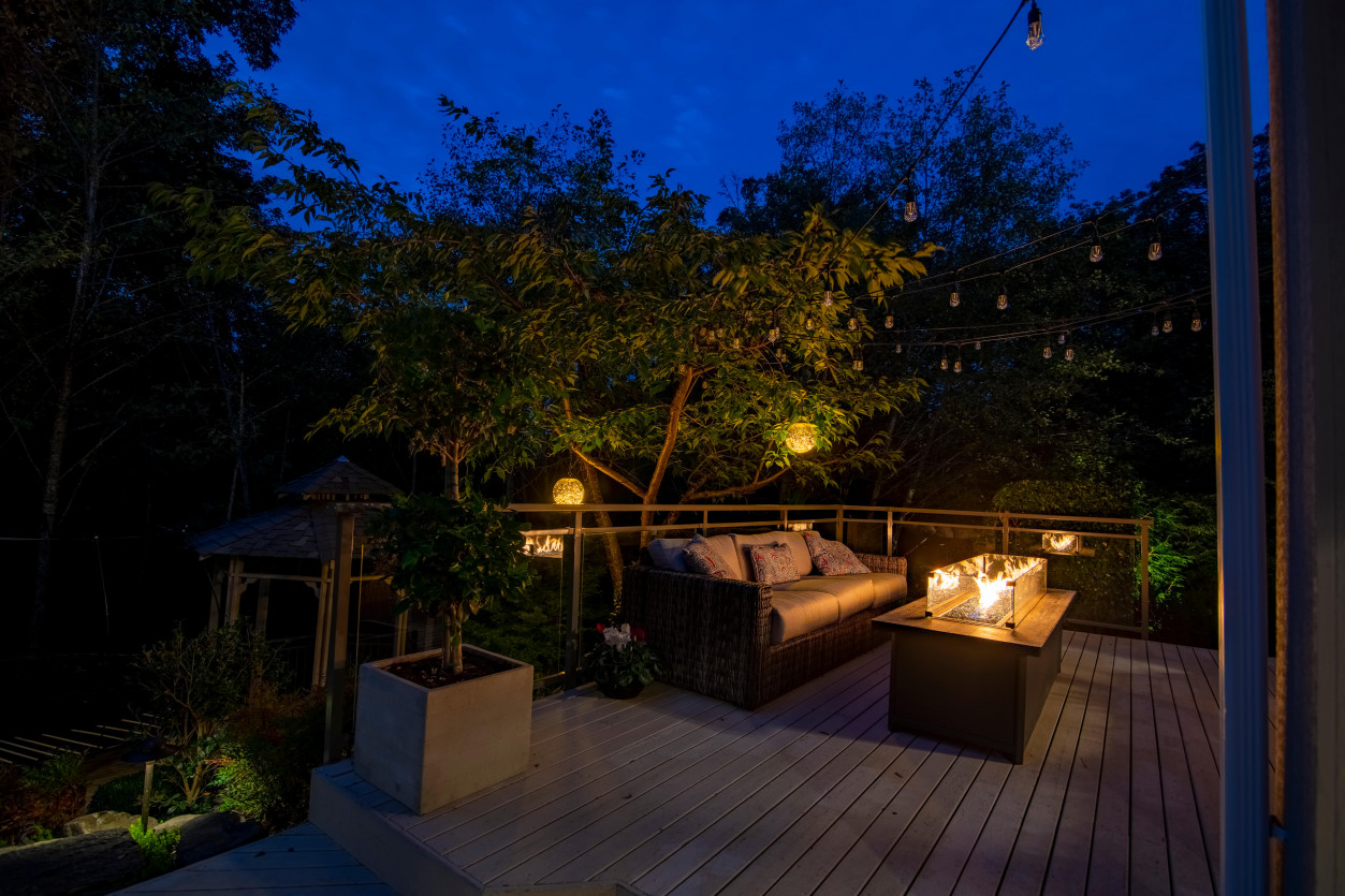 Nighttime view of illuminated firepit and deck seating overlooking wooded backyard.