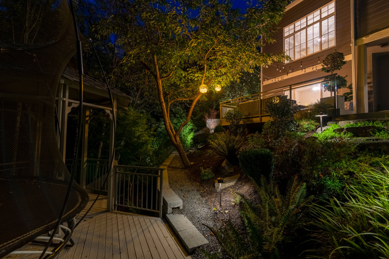 Multi-level deck in Sammamish illuminated with warm outdoor lighting, highlighting trees, lounge seating, and landscaped garden pathways.