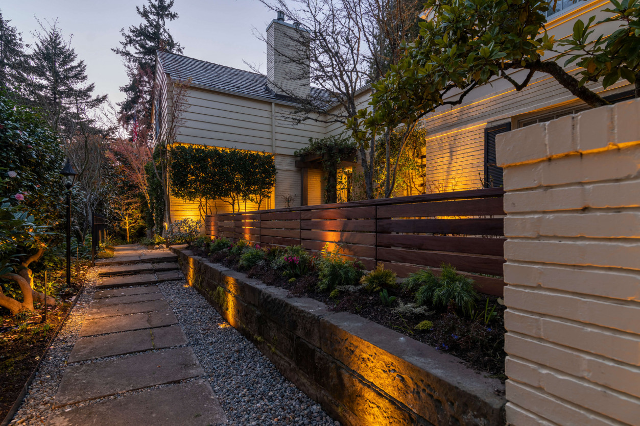 Architectural and pathway lighting along wooden siding and stone retaining wall in Magnolia backyard.