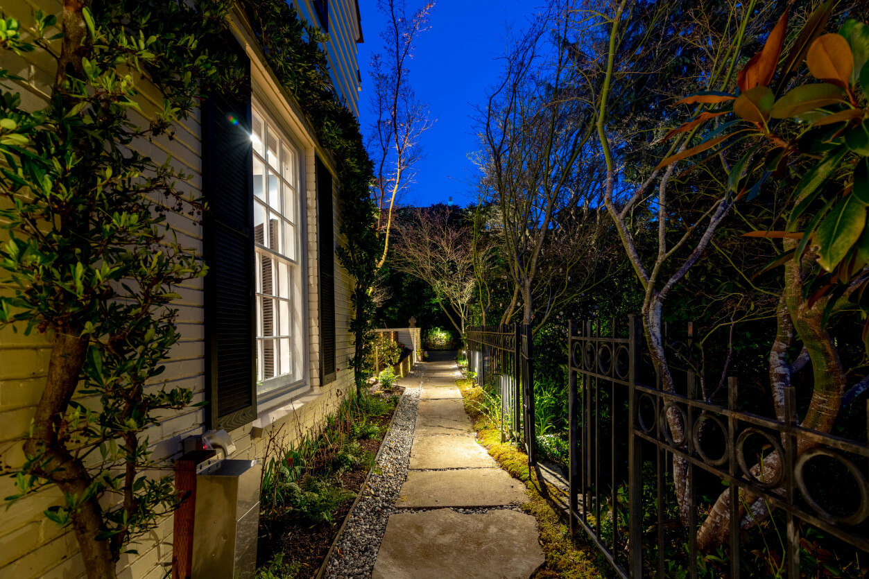 Narrow pathway lighting framed by tall fence and dense greenery with warm amber glow.